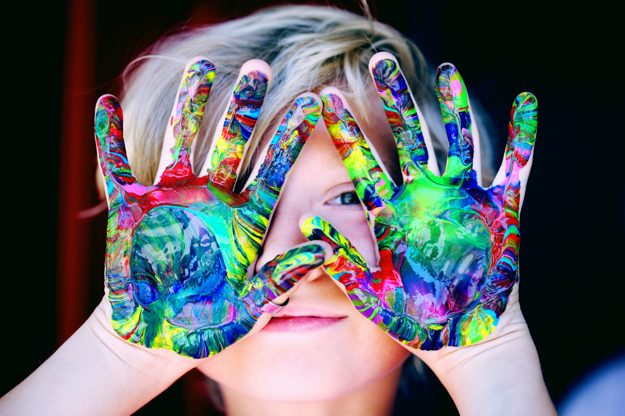 A child holding up their left hand over their face, with their palm facing the camera, with their hand covered in multiple colors of paint.