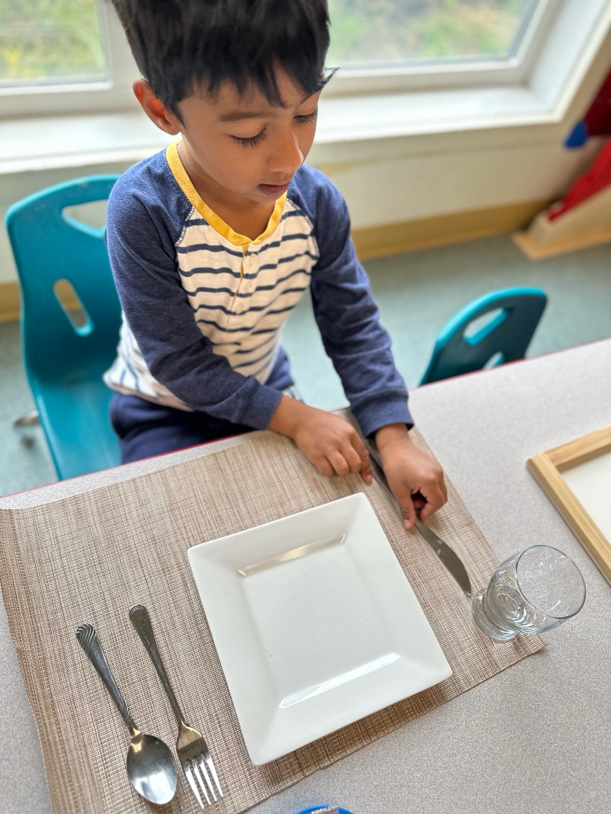 A boy practicing practical life skills by setting the table with cutlery, a plate and a glass.